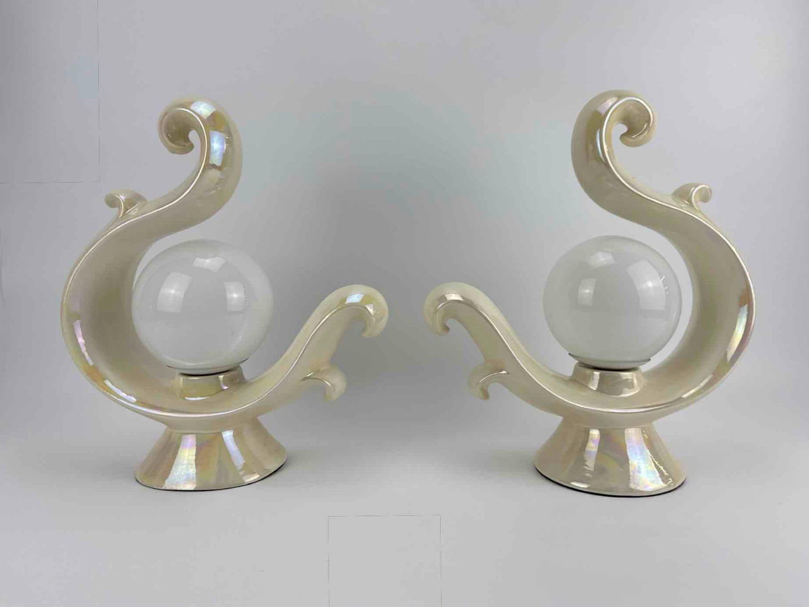 PAIR OF IRIDESCENT LUSTRE GLAZE CERAMIC SWAN FORM TABLE LAMPS WITH GLASS GLOBE SHADES: Pair of iridescent mother of pearl lustre glazed ceramic table lamps, stylized swan or scrolled wave form cradling a white glass globe shade, raised on flared circular ceramic bases Approximate Dimens