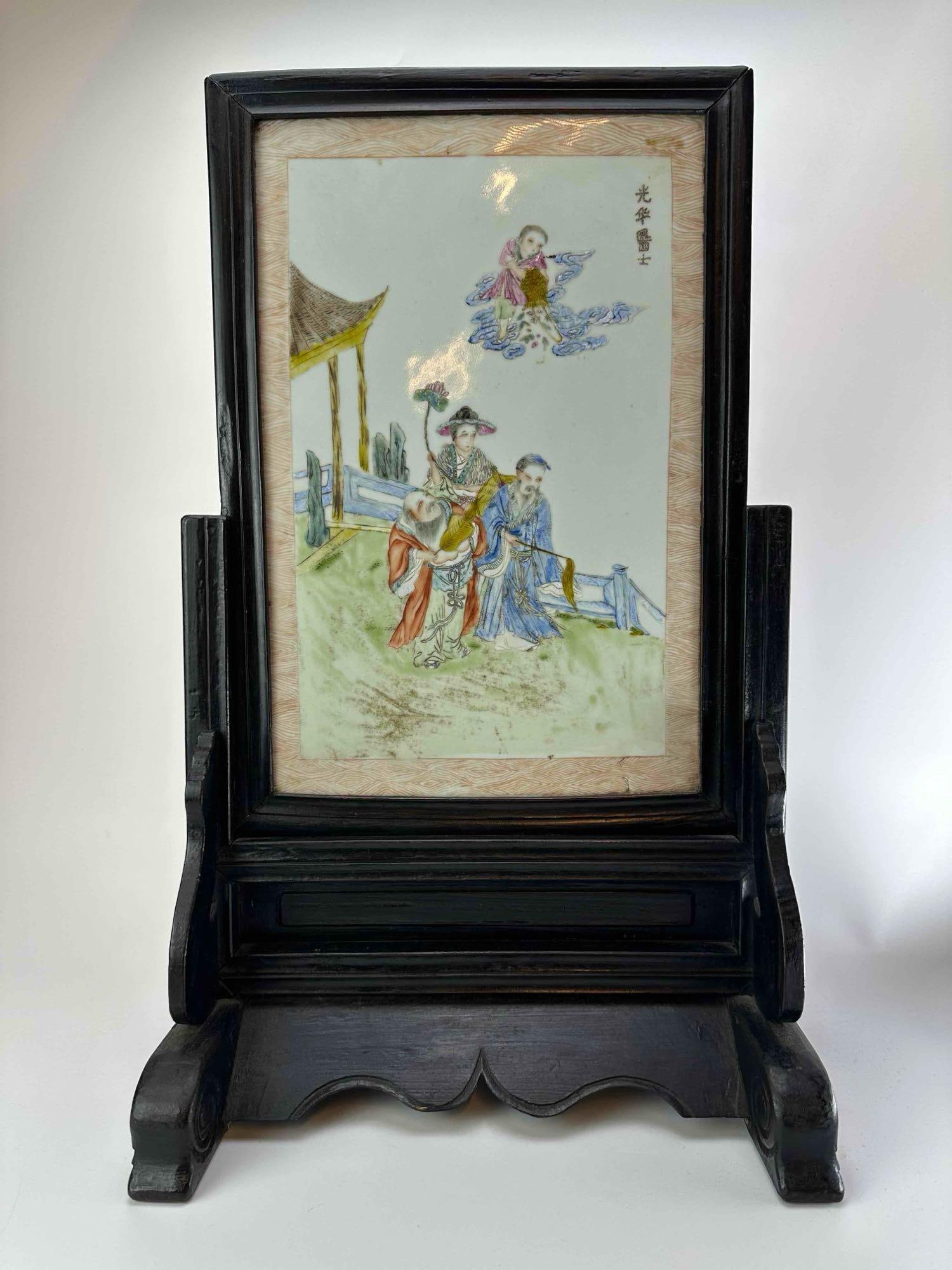 CHINESE HAND PAINTED PORCELAIN PLAQUE IN EBONIZED WOOD TABLE SCREEN STAND: Rectangular porcelain plaque painted in polychrome enamels with figures in a garden pavilion and an inscription, set in a carved ebonized wood table screen stand Approximate Dimensions: 23.5" H x 