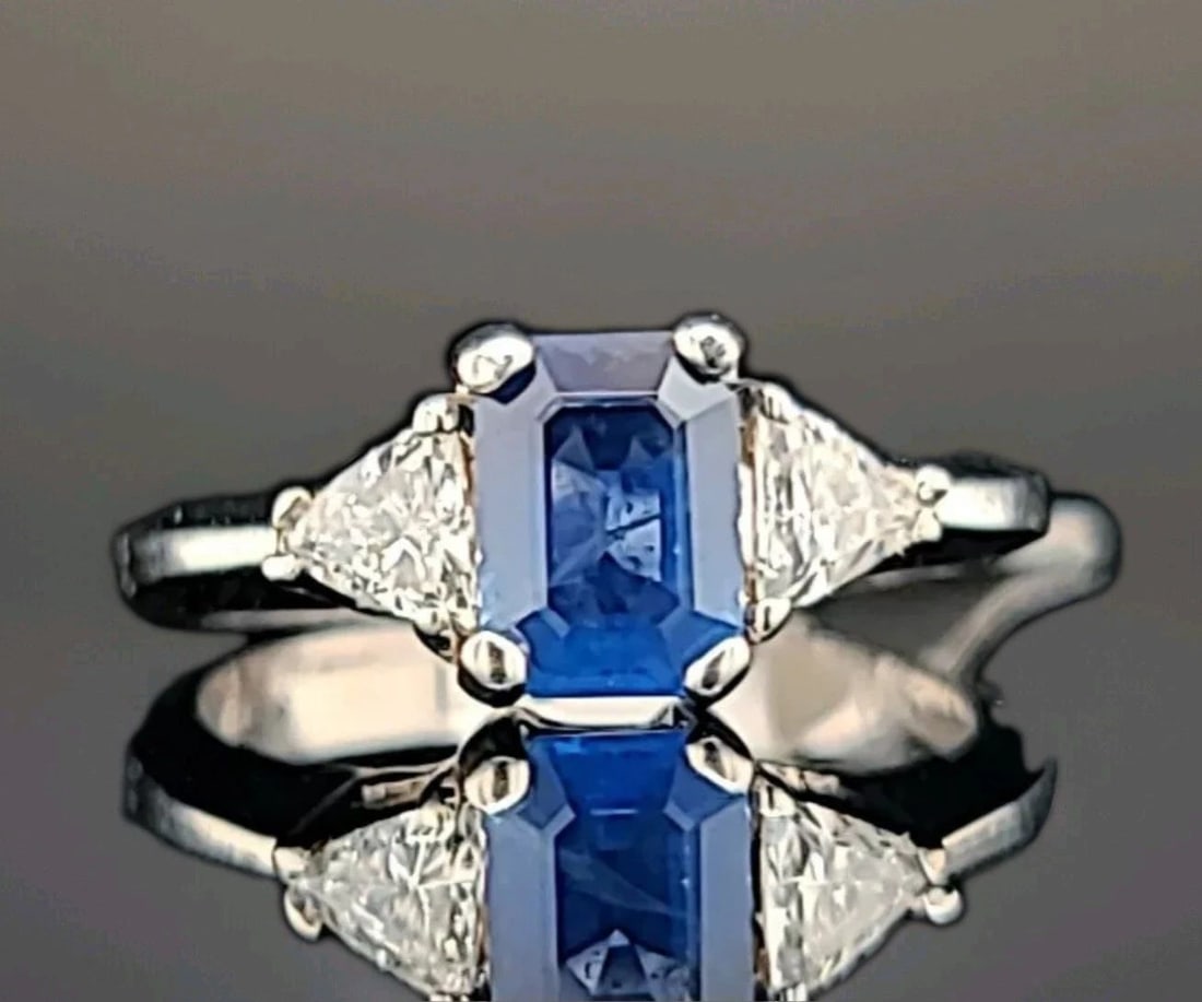 18K WHITE GOLD ENGAGEMENT RING 1.15CT. NATURAL BLUE SAPPHIRE EMERALD SHAPE: 18K WHITE GOLD ENGAGEMENT RING 1.15CT. NATURAL BLUE SAPPHIRE EMERALD SHAPE This exquisite 18K white gold ring features a stunning 1.15CT natural blue sapphire in an emerald shape, accented with two sp