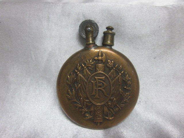 Round Brass Trench Art Petrol Lighter: On one side of the lighter, there is an angel and two horses that are engraved on it and on the other side it has FR in a circle on top of a scroll and axe head with flowers around it. I’m not sure
