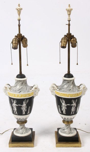 PAIR PAINTED CERAMIC BRASS VASE FORM LAMPS: A pair of painted ceramic and brass vase form table lamps, each having rams heads above classical scene of winged putti and urns. Ht: 33.25" Width: 9.5" Depth: 6.5"