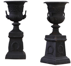 A CLASSICAL PAIR OF PAINTED CAST IRON GARDEN URNS RESTING ON A BASE WITH WREATH DECORATION.