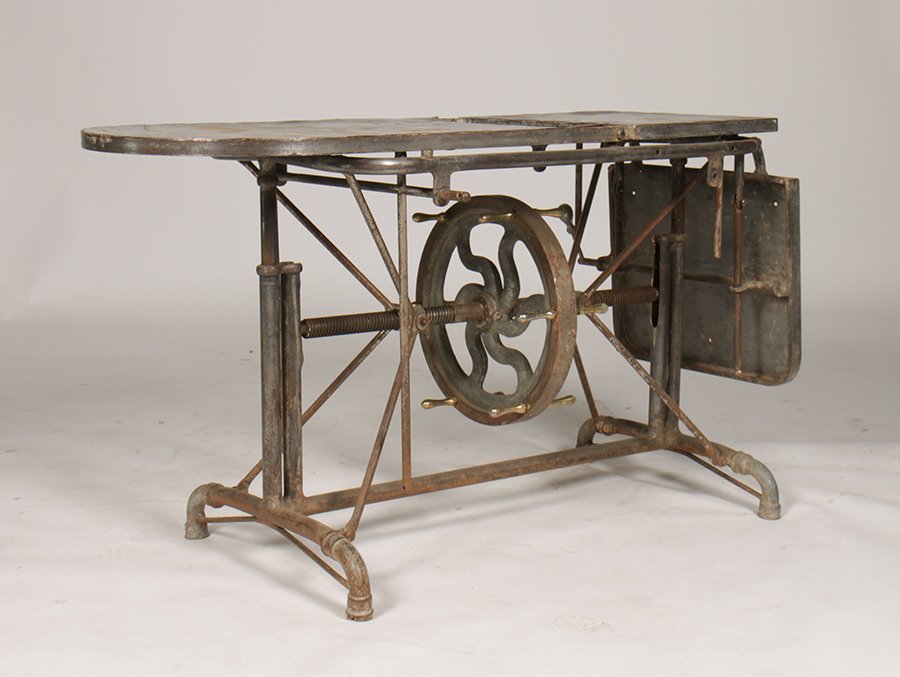 ADJUSTABLE MECHANICAL TABLE CRANK DRAWER 1900: Adjustable metal mechanical table having hinged top plate in three sections over a lower crank drawer wheel circa 1900. Raised on a trestle bottom. Ht: 29" Width: 61" Depth: 21"