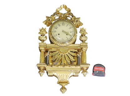 CARVED WOOD HANGING WALL CLOCK: Gilt. Mirrored back panel. Carved wood. White face with sunburst and flowers. 24 1/2" h x 12 3/4" w x 5" d.