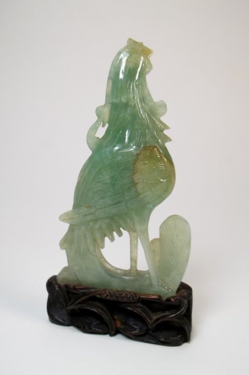 A Chinese Carved Jadeite Figure of a Phoenix: A Chinese jadeite carving, depicting a standing phoenix bird, with its head looking upward, one leg is stepping forward from another, its tail curved forward from the back toward to the middle of the 