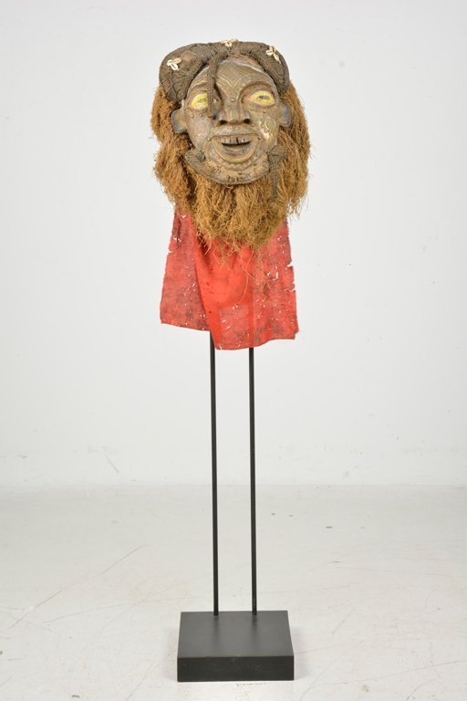 An African Carved and Beaded Wood Mask: Having applied metal and beaded decoration, with raffia hair and red cloth neck drape, and raised on the affixed metal and ebonized wood base.Size: 13" L x 10" W x 59 1/2" H (inclusive of base)