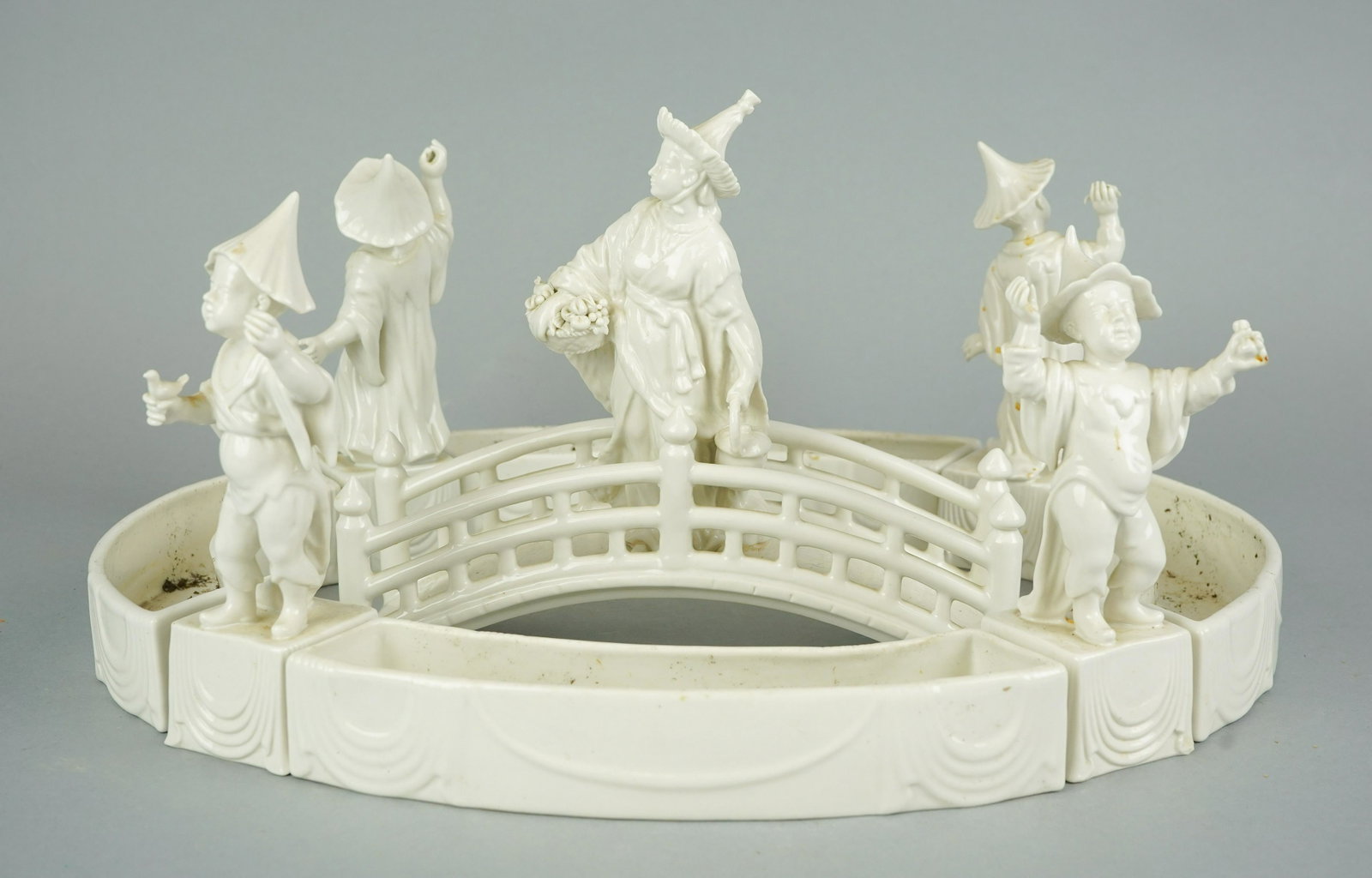 9-Piece Mottahedeh Design Figural Grouping: 9-Piece Mottahedeh Design blanc de chine centerpiece, Asian theme, flower ring with figures at corners, center figure on bridge (9" w x 8" h), overall approx 16-1/2" x 12", 1 figure with damaged hat, 