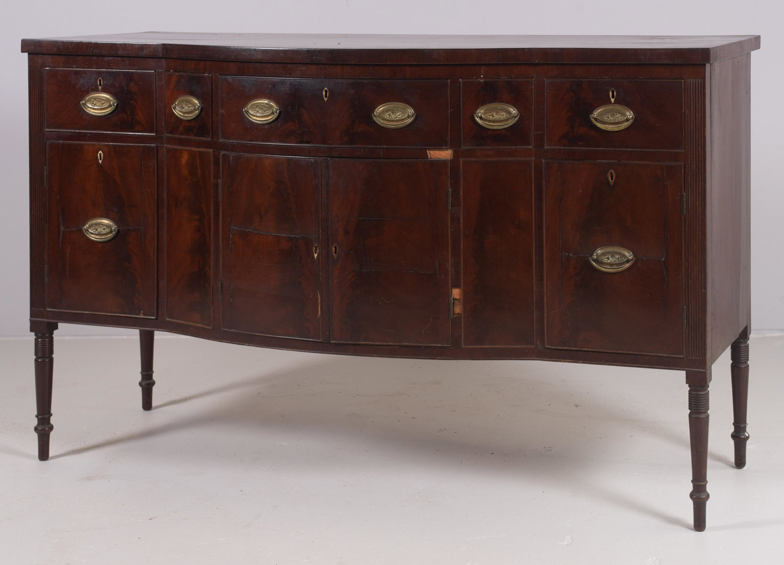 Mahogany Sheraton bowfront sideboard: Mahogany Sheraton bowfront sideboard, frieze with five drawers over conforming doors, replaced brasses, all flanked by reeded styles, raised on turned and reeded legs, 66"w x 24"d x 40"h, Philadelphia