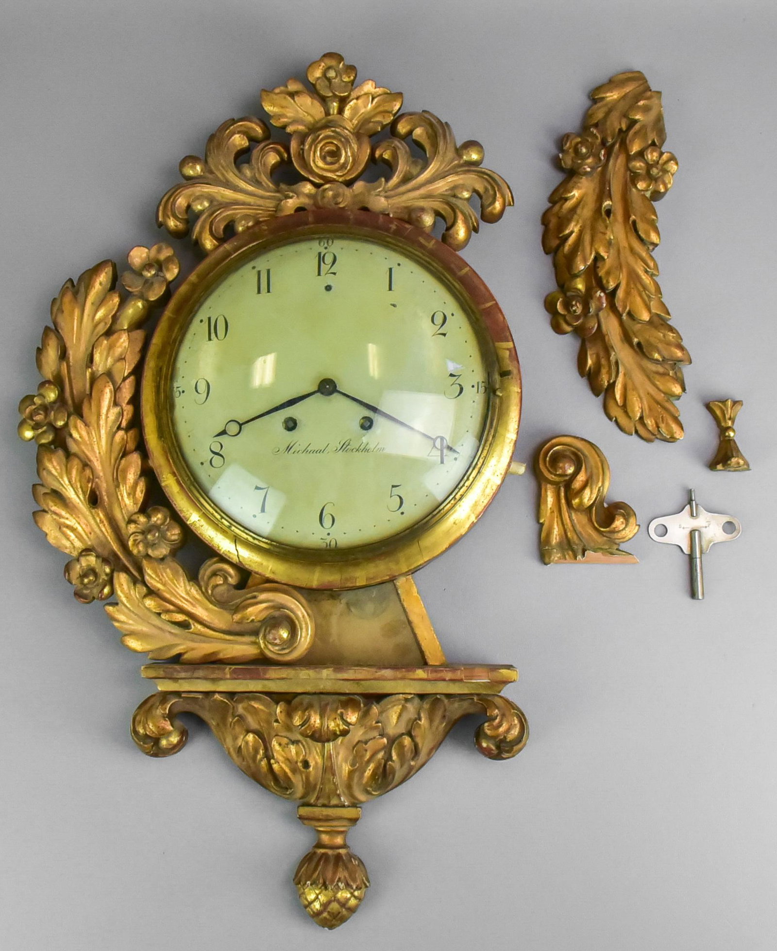 Swedish Carved Wood Wall Clock: Swedish carved and gilt painted wood wall clock, 9" dia enamel dial marked "Michaal, Stockholm", with key and pendulum, 26" h, right side foliage needs to be repaired & reattached, missing rose at top