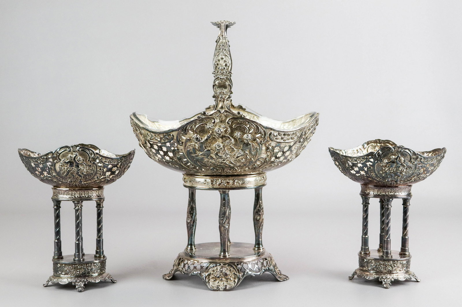 3-Piece Silver Plate Compote Set: 3-Piece silver plate compote set, reticulated with embossed cherubs, c/o 19-7/8" x 7-1/4" x 13-1/2" h basket with contoured glass insert, (2) 6" x 4-5/8" x 7" h compotes (missing inserts)