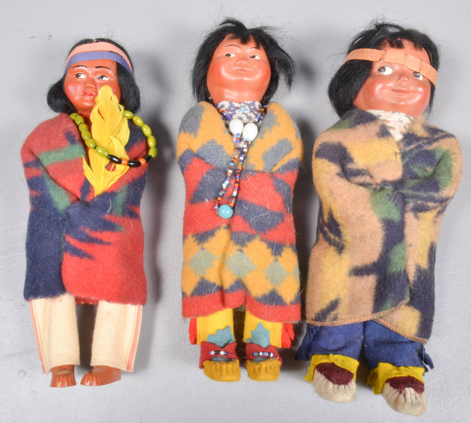 (3) Skookum Dolls: (3) Skookum dolls to include squaw baby, 9", plastic feet, no label, green beads and feather, young boy, 9", felt feet, red, blue and yellow with no label and young boy, 9" felt feet with white, yello