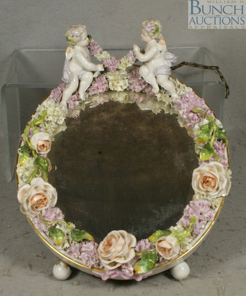 Oval German porcelain beveled dresser mirror wit: Oval German porcelain beveled dresser mirror with applied floral border and winged cherub crest, usual losses to flowers and foliage, one cherub hand with minor loss, 13" h, 10 1/2" w, unmarked