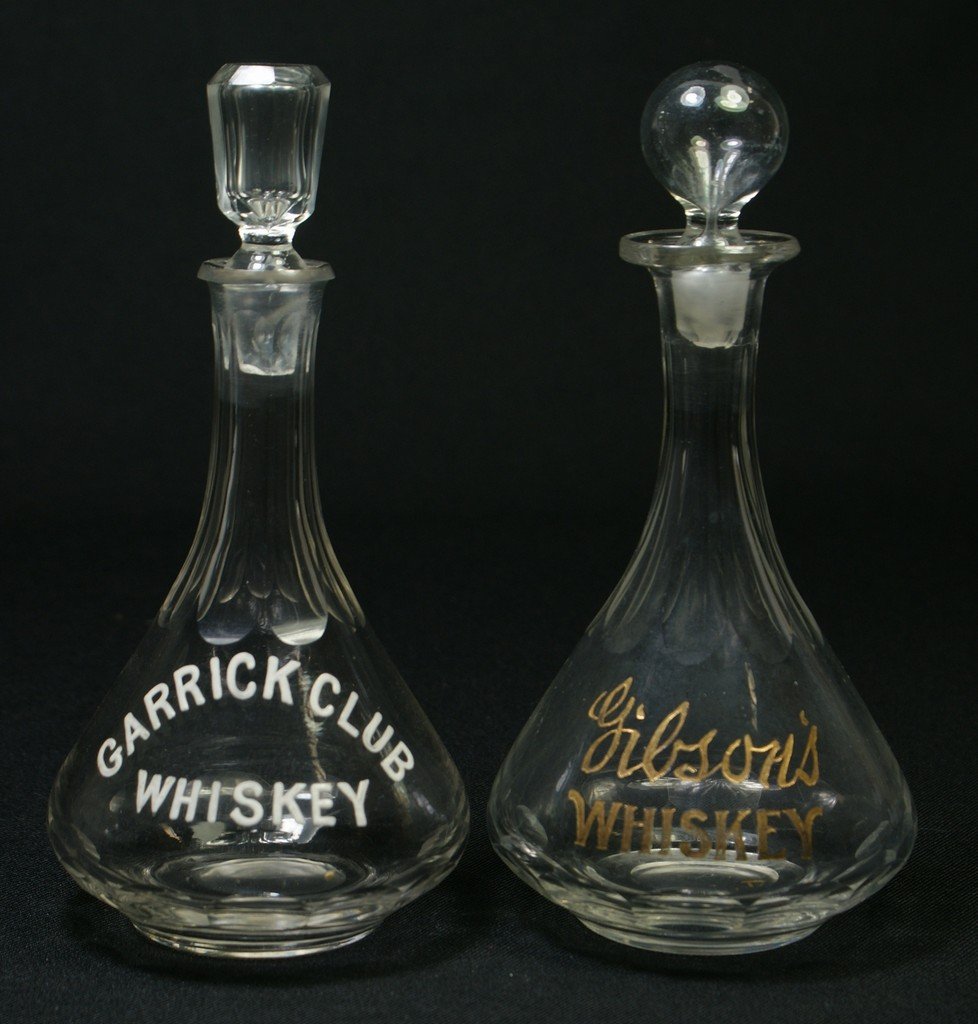 2 clear glass back bar bottles with etched, enam: 2 clear glass back bar bottles with etched, enameled, and gilt decoration, GIBSONS WHISKEY, GARRICK CLUB WHISKEY, tallest 8", chippage on bottom of one stopper