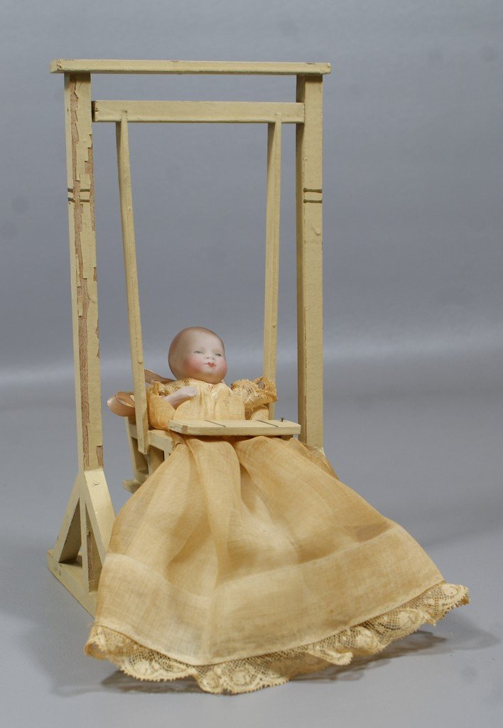 Tiny All Bisque Bye-lo with stationary neck, painted ey: Tiny All Bisque Bye-lo with stationary neck, painted eyes, molded socks and pink painted shoes, bearing the original paper label on torso, wearing original dress and sitting in a wooden swing rocker, 