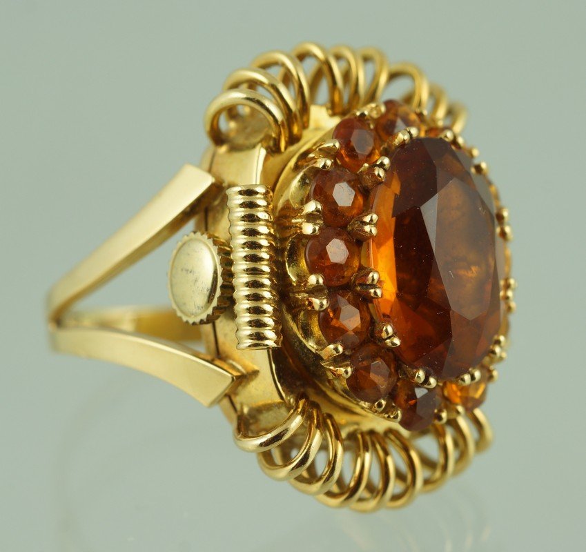 18 K YG watch ring, hinged top set with oval cut topaz: 18 K YG watch ring, hinged top set with an oval cut topaz, surrounded by 12 round cut pink stones, the dial of the watch marked Emka, Geneve, Incabloc, 8.9 DWT gross, size 3 3/4.