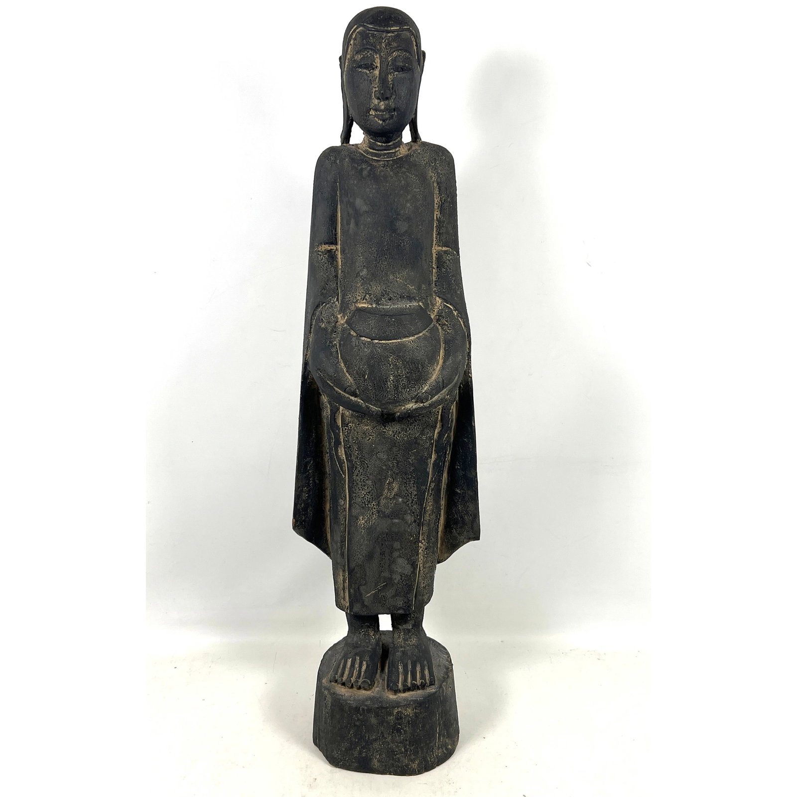Southeast Asian carved wood figure of a standing Buddha, depicted in a serene posture holding an alm: Southeast Asian carved wood figure of a standing Buddha, depicted in a serene posture holding an alms bowl with both hands. Dimensions: Height: 35. inches, Width: 6.75 inches, Depth: 6.5 inches. ---
