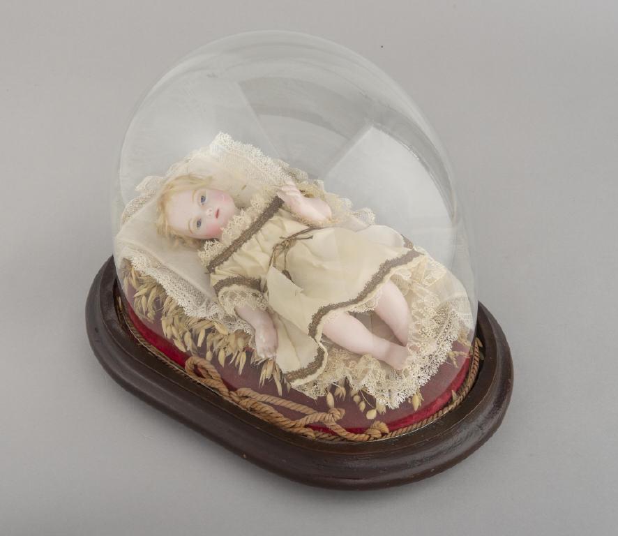 UNUSUAL WAX DOLL, CIRCA 1880: An unusual wax doll lying on a bed of wheat. Wax face, arms and legs, dressed in period dress and undergarments. Resting under a glass dome. Circa 1880. Dimensions, 12" x 14" x 10".