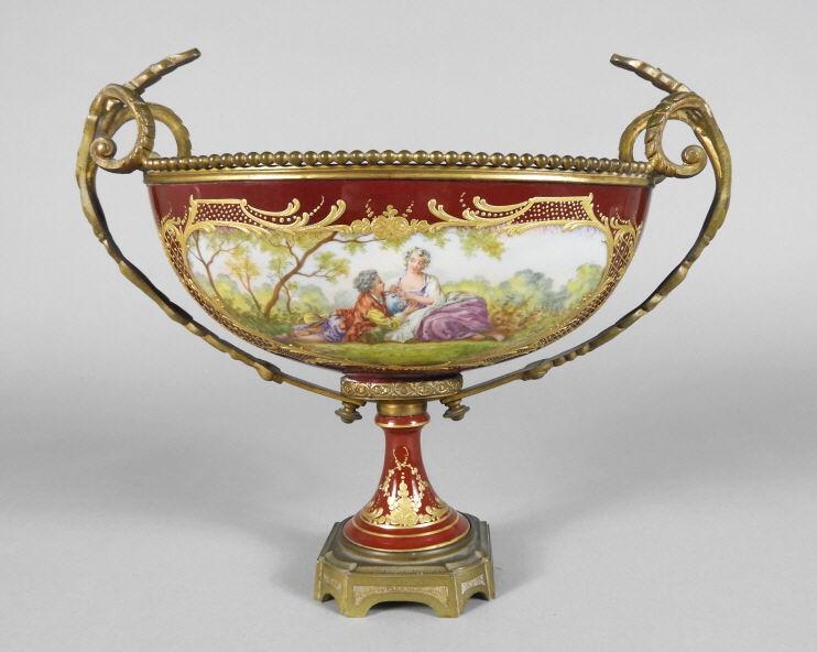FRENCH PORCELAIN COMPORT, LATE 19TH CENTURY: A late 19th century French porcelain comport, the oval body decorated with gilt detail and hand painted romantic vignettes signed 'Verjot' over a ruby red field, mounted within an ormolu frame raised 