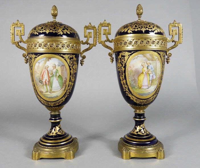 PAIR OF SEVRES STYLE LIDDED URNS: Pair of early 20th century Sevres style porcelain urns, both cobalt bodies decorated with hand painted romantic vignettes signed 'Robin' and gilded classical motifs, fitted with ormolu mounts and rais