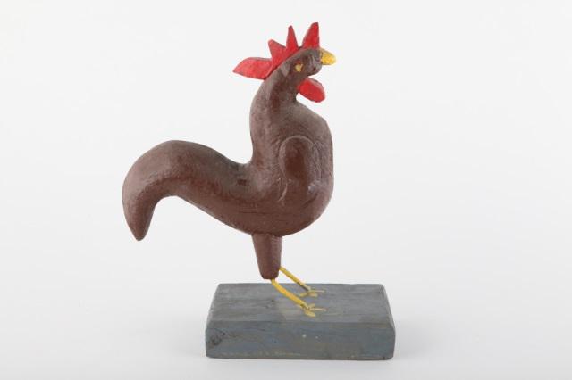 Naive Rooster Sculpture, Pierre Bisson, Circa 1980: A naive rooster sculpture in brown paint with iron rod legs on a grey base, carved by Pierre Bisson, Quebec, circa 1980. H: 16"; W: 13"; D: 6".