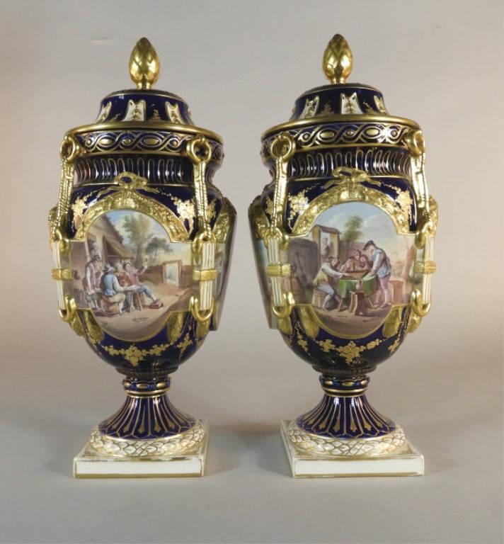 Pair of Continental Glazed Earthenware Vases: A decorative pair of early 20th century Continental 'Sevres-style' glazed earthenware vases, both cobalt lidded bodies decorated overall with gilt detail and four painted panels of rural themes signed