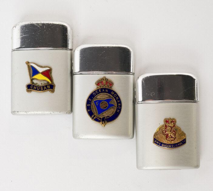 Vintage Passenger Ship's Souvenir Lighters: A group of three vintage Ronson flip-top passenger ship's souvenir cigarette lighters, the matte cases decorated with enameled cartouches. Each measuring 2.25" high.