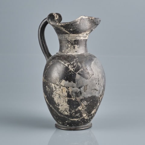 ANCIENT GREEK OINOCHOE: Greece, circa 300 BC, the black glazed pyriform earthenware wine jug body rising to a waisted neck with incised linear decoration, to a trefoil spout affixed with an integral applied handle, raised ov