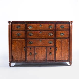 VERY GOOD SHERATON INSPIRED OAK SIDEBOARD: Northeast Atlantic, circa 1830, the figured top with porringer corners over full length reeded pilasters, fronted by a series of drawers, cupboards and twin cellarettes, all with beaded bird's eye map