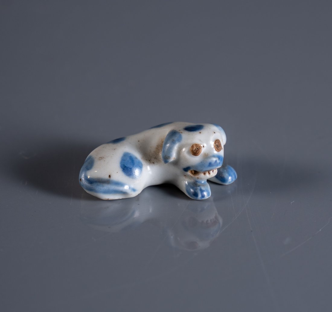 HIRADO PORCELAIN NETSUKE OF A PUG: Japan circa 1850, the recumbent white porcelain figure of a pug dog shown baring its teeth, decorated with spot painted cobalt detail, the underside with twin holes intended for a cord to pass through
