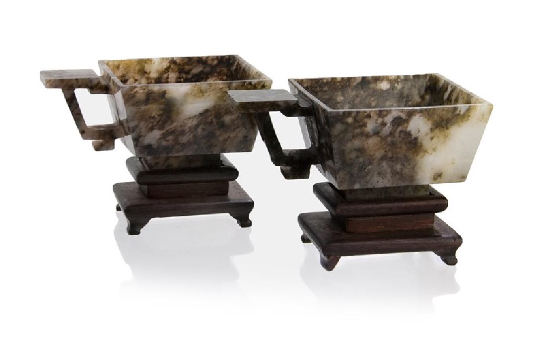 CARVED JADE CUPS ON STANDS: Of rectangular form on an extended foot from a translucent jade mottled with dark brownish green inclusions. _x000D_ H:1 1/2in(3.8cm) W:3 1/4in(8.3cm) M:2 1/2in(6.4cm) 153g