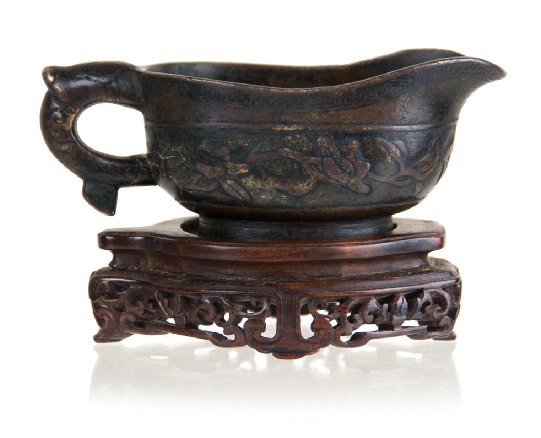 MING DYNASTY BRONZE VESSEL ON BASE: A Ming dynasty bronze vessel molded with an elephant head handle, floral patterns on the body, and an extended mouth. Wooden base included. H:1 1/2in(3.8cm) W:4 1/4in(10.7cm)