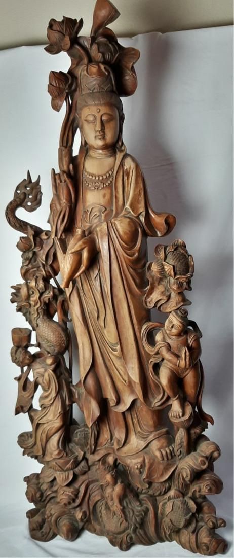 A DELICATE WOOD CARVED FIGURE OF GUANYIN: Deftly carved Guanyin figure dressed elegantly in long flowing robes standing on cresting waves, her face with gentle smile and her hair piled up into a high chignon, holding a flowering lotus stem on