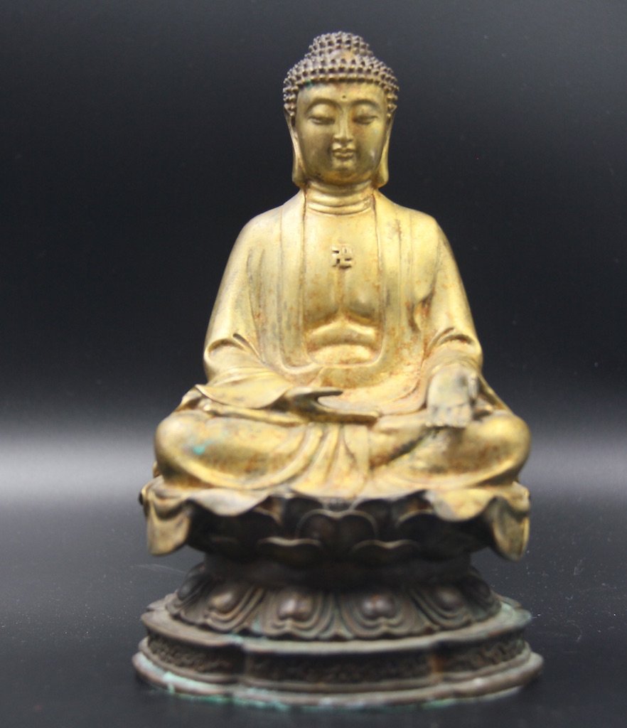 GILT-BRONZE BUDDHA: The figure is shown seated in dhyanasana with hands in dhyana mudra, and wearing simple draping robes falling in graceful folds that expose a bare chest that bears a swastika. The face is well cast in