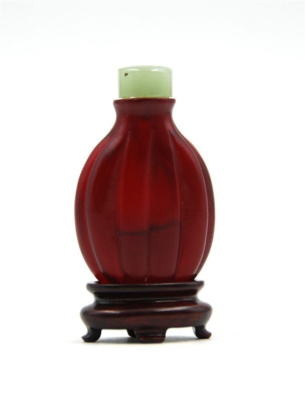 RIDGED GLASS SNUFF BOTTLE: This ruby red ovoid bottle is marked with ridges that taper off near the neck and rests upon a wood stand shaped like an incense burner. Topped with a pale jade stopper. H: 2 1/2