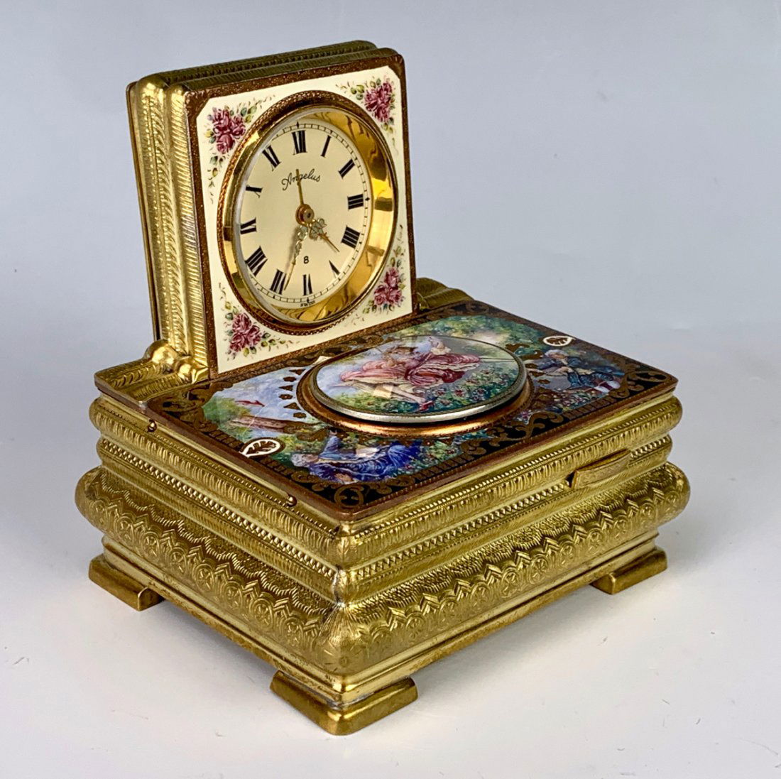 Rare Greisbaum Enameled Singing Bird Box with Clock: First half of the 20th century, square clock with an Angelus wind up alarm movement, mounted on a gilt metal and enameled bird box, painted to depict a scene of a woman in a swing, the pop up oval ope