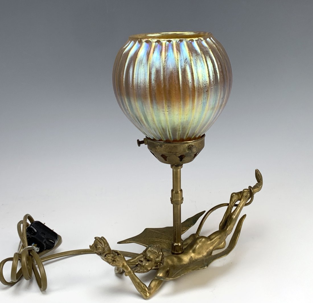 Gilt Bronze Devil Blowing Horn With A Favrile Shad: Quite unusual! Having a devil with wings laying down and blowing a horn. His tail is wrapped around his feet. The shade is a ribbed iridescent gold glass. Electrified with a switch. Circa 1910. Size: 