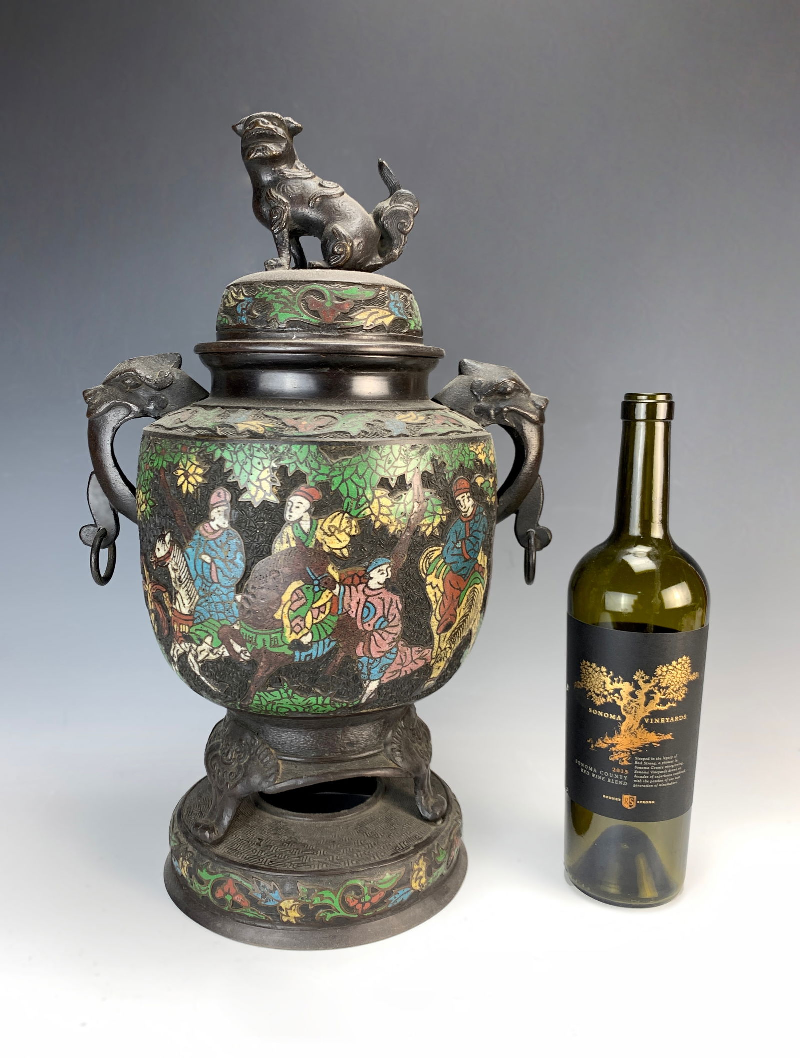 19th C Japanese Champleve Enamel Covered Urn: Meiji period, Japanese, late 19th century., bronze body with enameled (champlevé) scenes of warriors on horses, featuring two grotesque heads as handles and a Foo dog bronze figure at the top. Signed