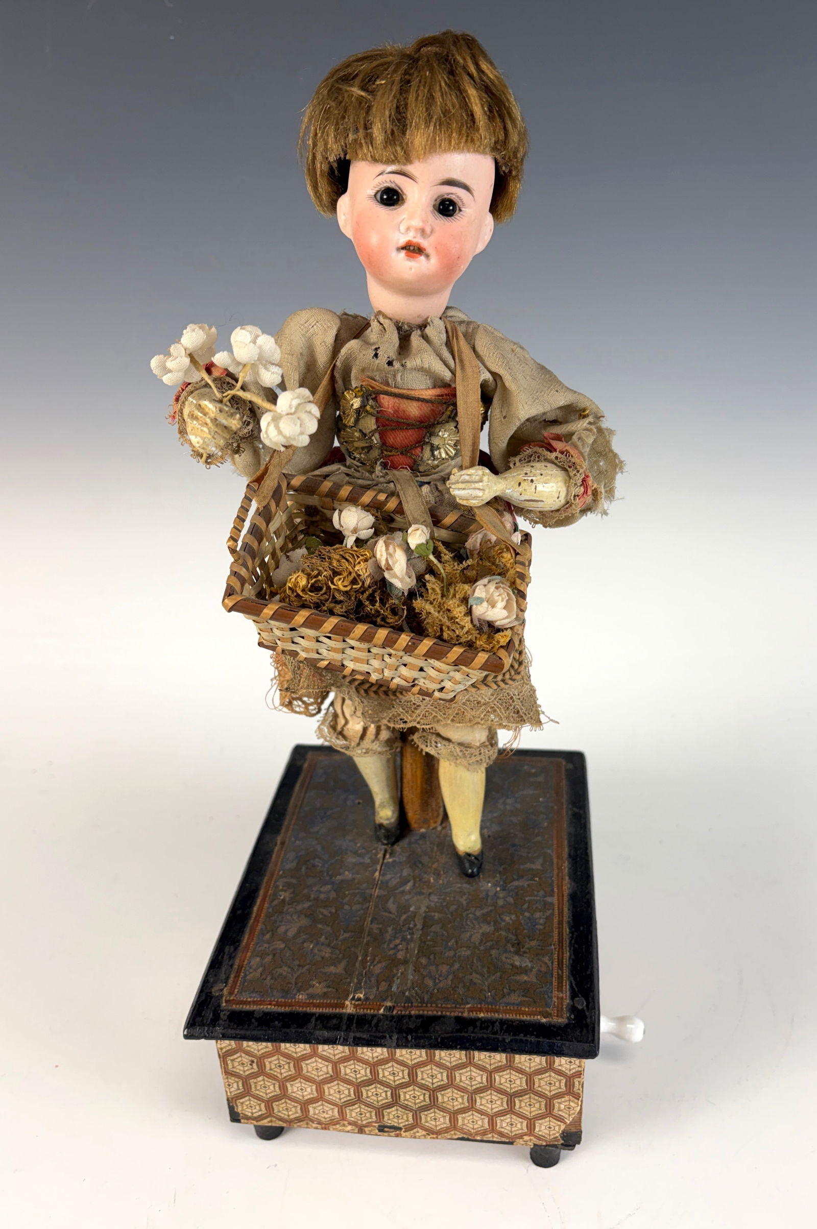 Antique Bisque Head Doll “Flower Seller?? Automaton: A bisque head doll automaton depicting a “Flower Seller,” circa 1900. When the handle is wound, she moves her head side to side and her right arm sways back and forth, accompanied by music. 13