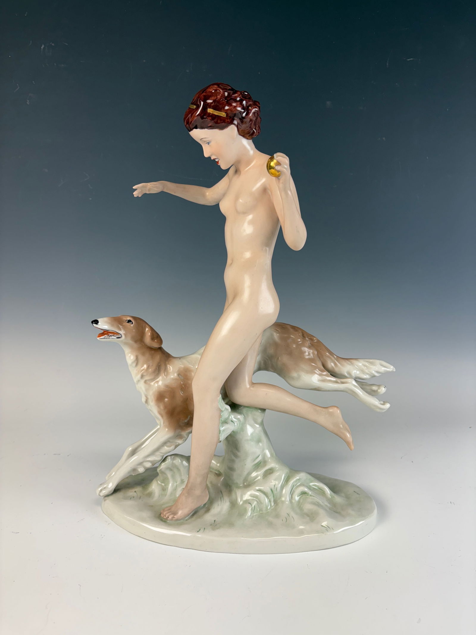 Royal Dux Porcelain Figure of a Nude Woman & Dog: A fine porcelain figure depicting a nude woman holding a gold ball with a running dog at her side, made by and signed Royal Dux. 14.25” tall × 11” wide × 6” deep. In excellent cond