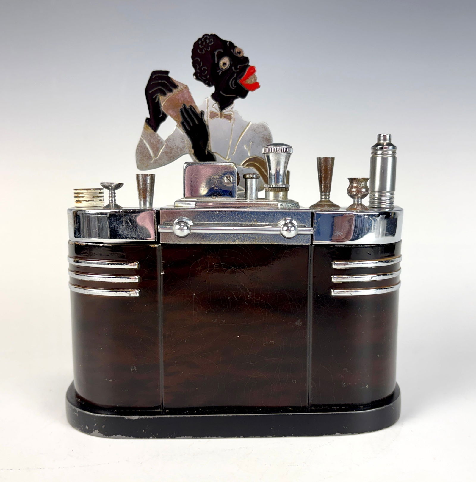 Black Americana Ronson Bartender Lighter: A decorative Ronson lighter in Black Americana style, featuring a chrome and enamel woodgrain-effect bar with a touch-tip lighter and two hinged cigarette compartments. Measurements: 6.75” tall