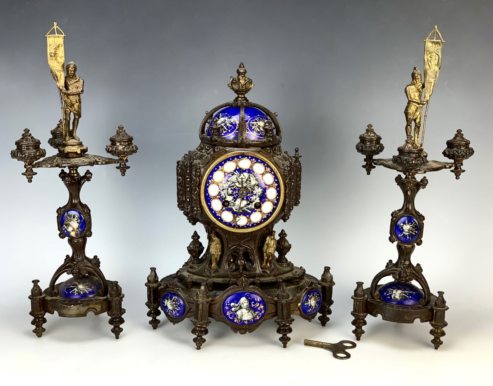 Patinated Metal & Enamel 3pc. Clock Garniture: Circa 1880, French ornate metal clock set with figures and blue enameled scenes, each matching candelabra has gilt metal figures with knights holding flags, good condition, 15" candelabra height *Cloc
