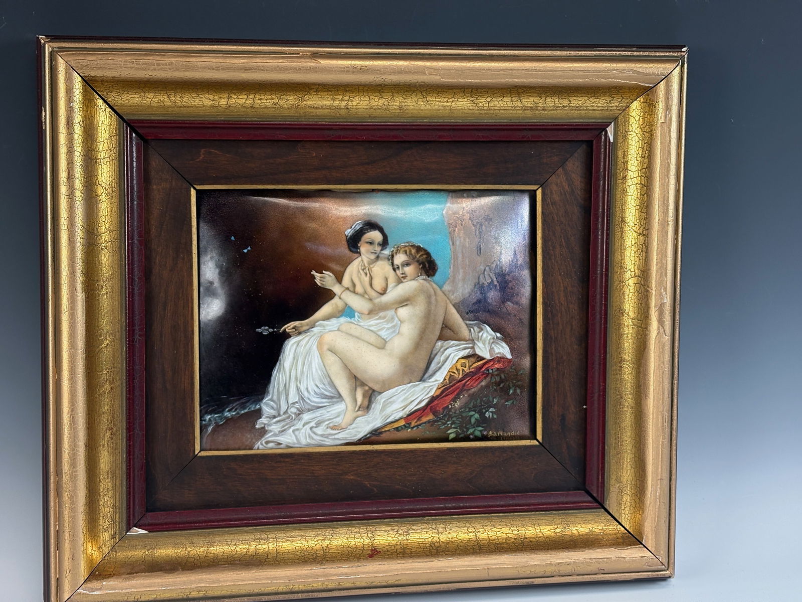 Sarlandie “Two Nudes?? Enamel Plaque: An enamel plaque by Sarlandie depicting two nude women seated on a rocky knoll with mountains in the background, finely rendered with rich color and detail. In very good condition; the frame has a cou