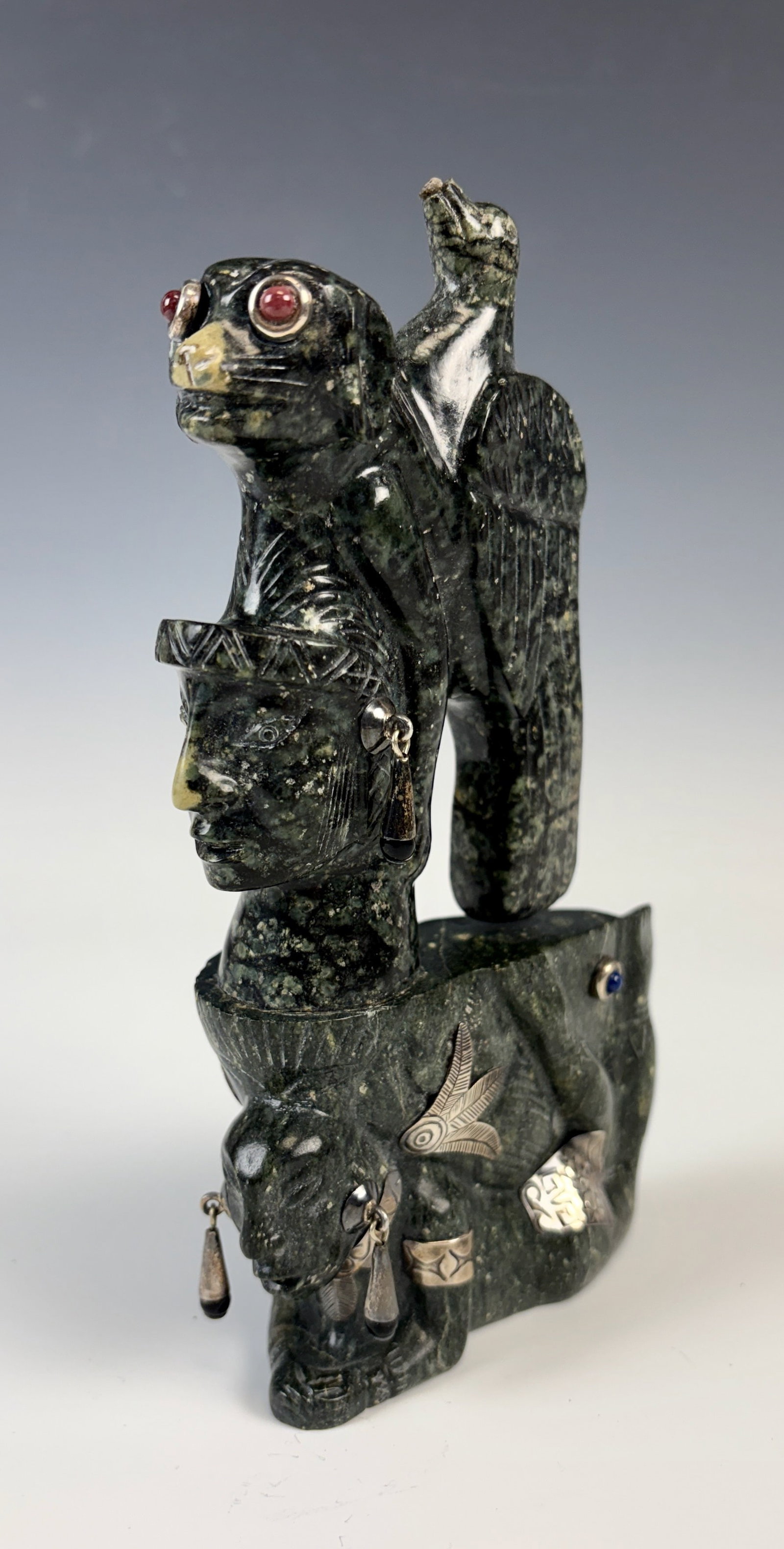 Carved Stone Aztec Figure w Silver Adornments: Dark green carved stone figure having a mans face above with is a dog head and a bird, with silver accents and other attachments including a cuff bracelet and earrings, with cabochon accents, very goo