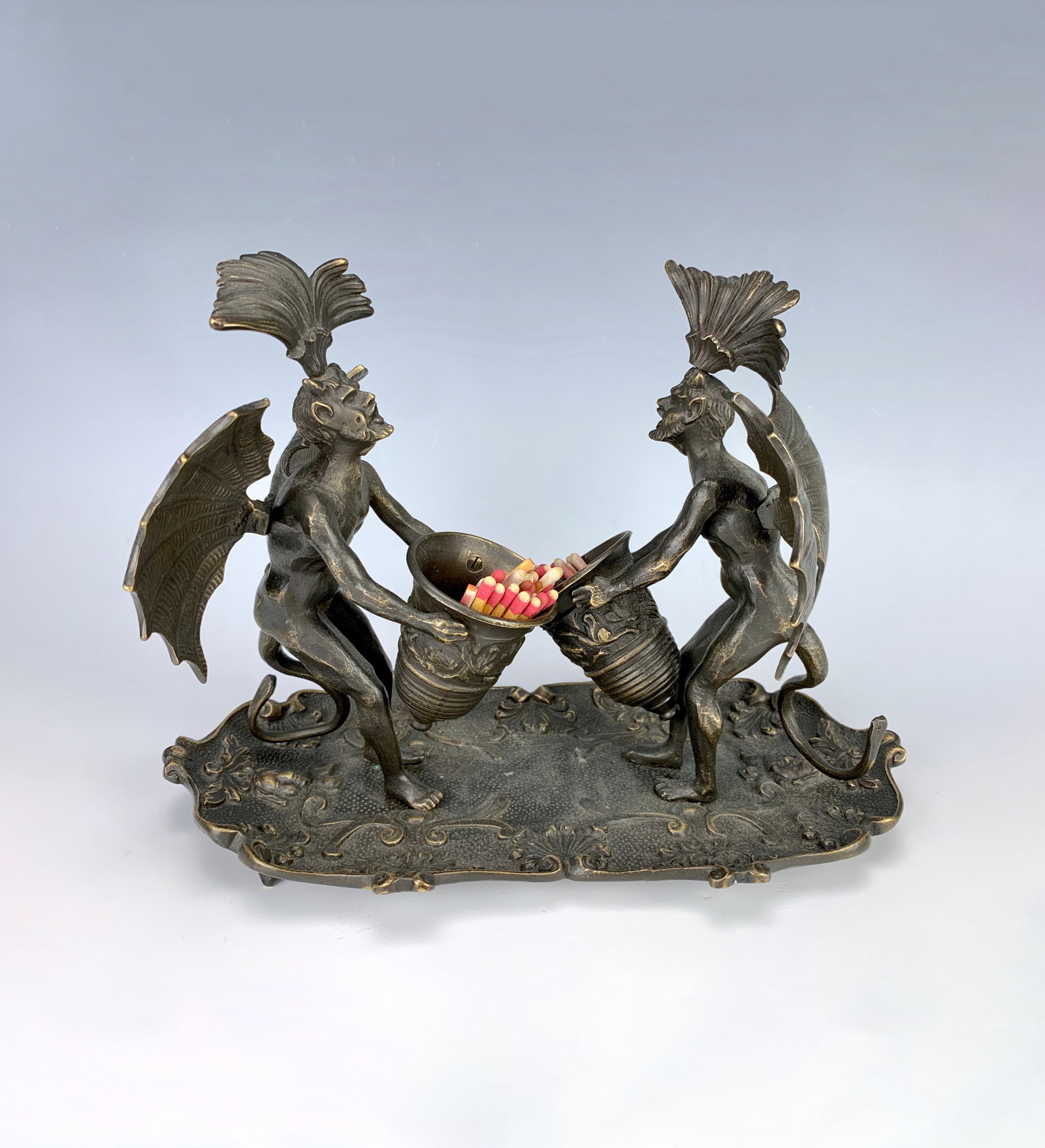 Rare Antique Bronze "2 Devils" Match Holder C.1880: Rare tobacciana item formed as 2 winged bronze devils holding urn shaped match holders, mounted on a bronze cartouche shaped foliate scroll and mask heads, very good condition, 7.5" x 10" x