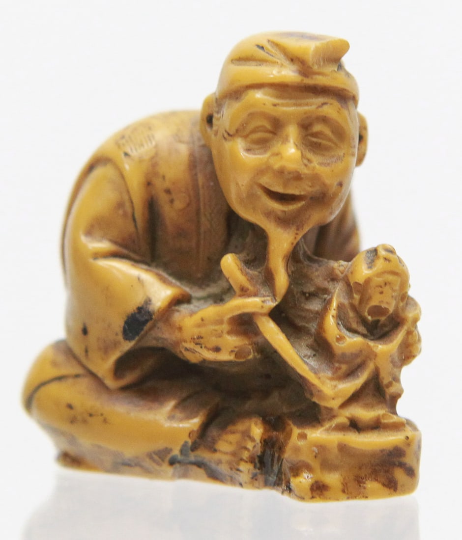 Japanese Carved Netsuke of Elderly Man Holding Child Figure: Finely carved Japanese netsuke depicting a seated elderly man with a smiling expression, long beard, and traditional robe while holding a small child or doll figure in his hands. The carving features 