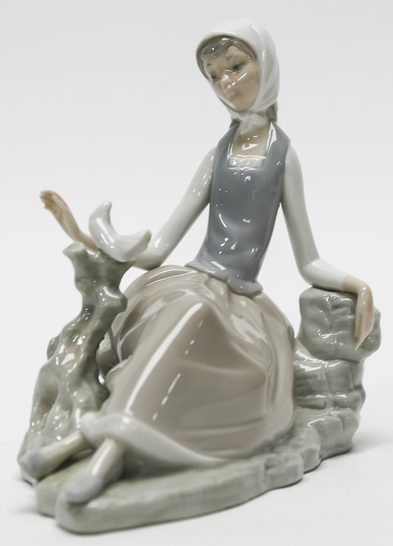 Lladró Shepherdess with Dove Porcelain Figurine #4660: Lladró glazed porcelain figurine depicting a graceful seated shepherdess wearing a headscarf, gray bodice, and flowing taupe skirt as she reaches toward a white dove perched on a sculpted tree