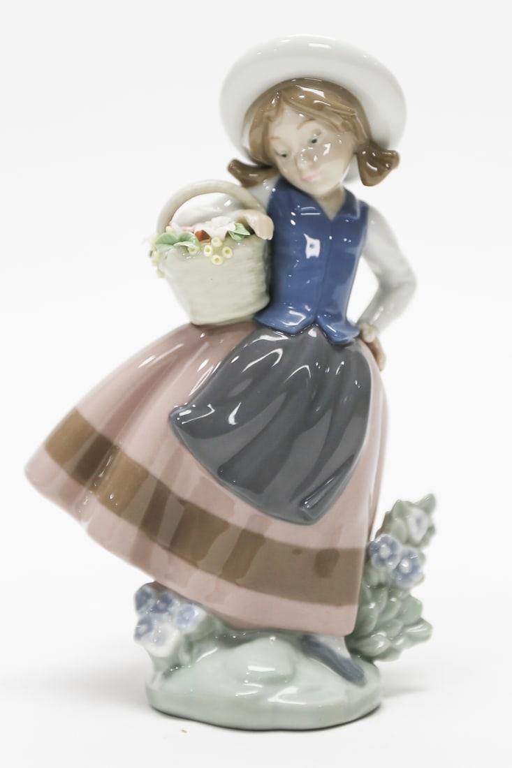 Title: Lladró Flower Girl Figurine: Description: Lladró glazed porcelain figurine depicting a young flower girl wearing a white hat, blue vest, gray apron, and flowing pink skirt with brown trim. She carries a woven basket filled with