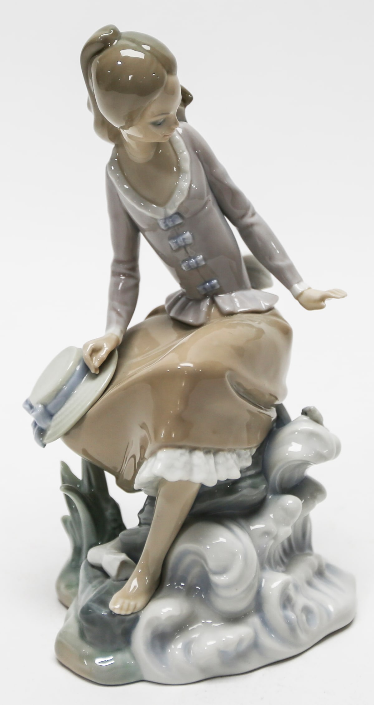 Lladró Seated Lady by the Sea Figurine: Elegant Lladró porcelain figurine depicting a young woman seated on seaside rocks beside a large conch shell. She wears a soft lavender jacket with blue bow accents, a flowing brown skirt, and holds 