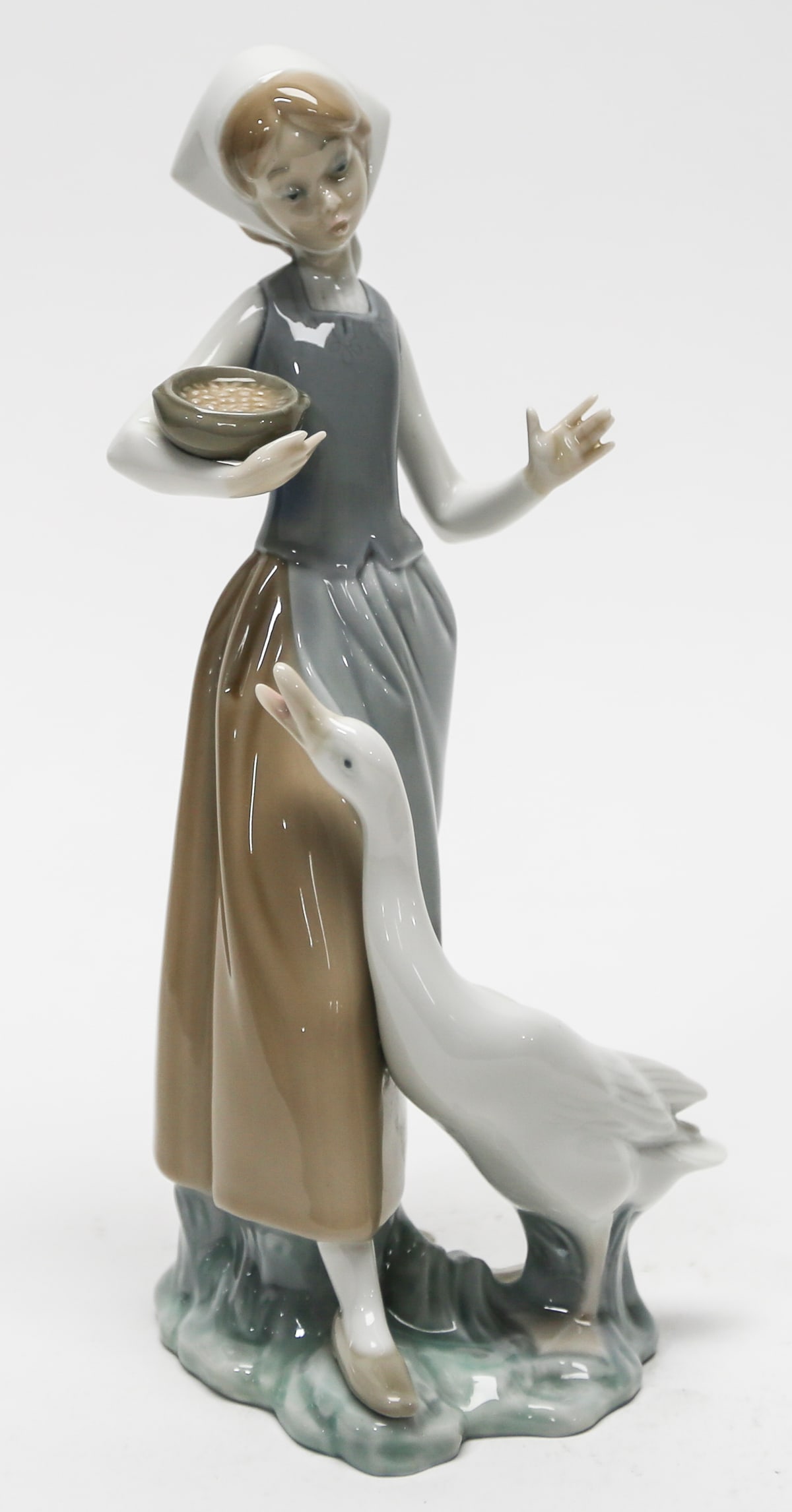 Lladró Goose Girl Porcelain Figurine: Charming Lladró porcelain figurine depicting a young farm girl in a white headscarf and apron carrying a bowl of grain while a tall goose stands attentively beside her. Rendered in Lladró’s