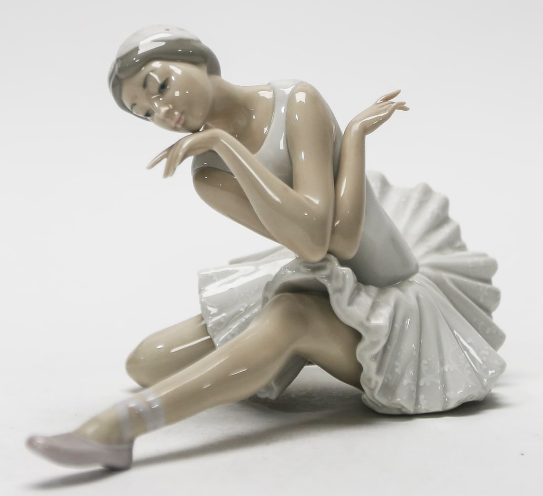 Lladró Seated Ballerina Porcelain Figurine: Elegant Lladró porcelain figurine depicting a seated ballerina in a graceful resting pose, dressed in a finely detailed white tutu with pale lavender slippers. The sculptural composition captures